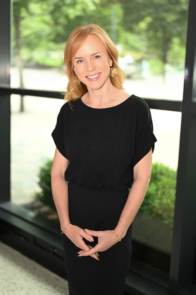 A smiling woman with blonde hair and blue eyes stands in front of a blurred green background.