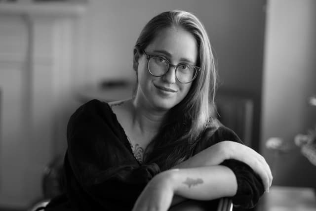 A woman with long blonde hair and glasses, wearing a black sweater, is smiling and looking directly at the camera in a dimly lit room.