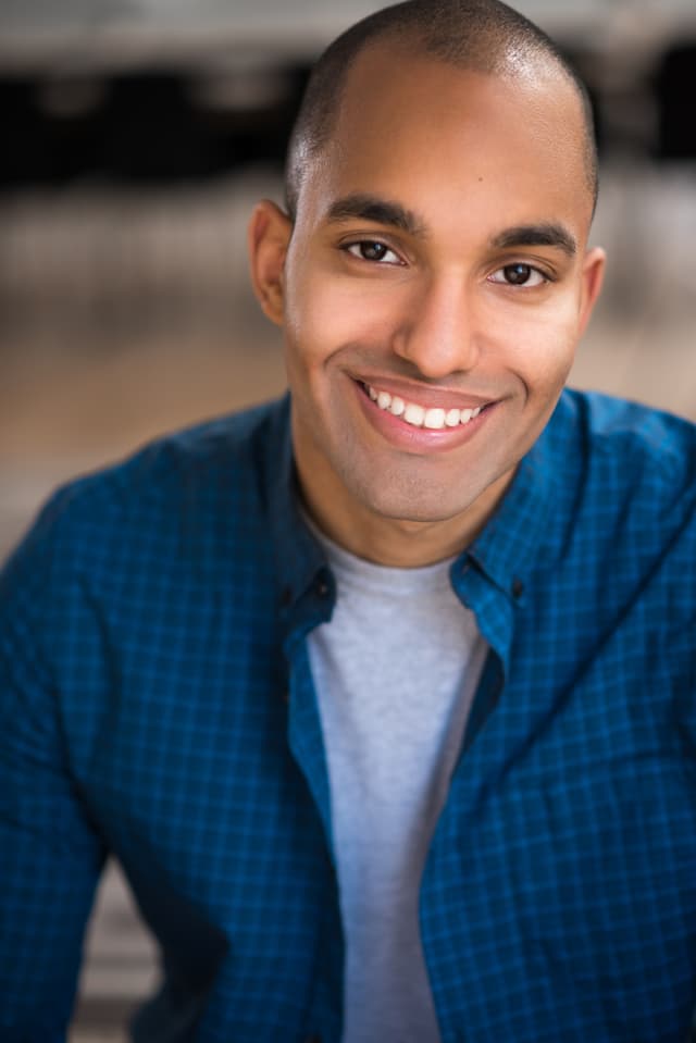 The image shows a smiling man with a bald head wearing a blue checkered shirt, set against a blurred background.
