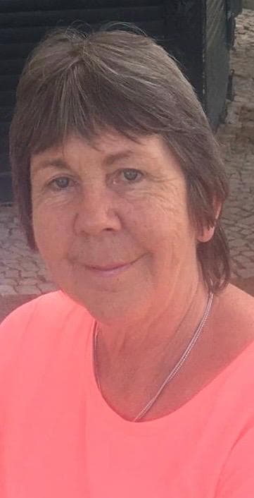 The image shows a middle-aged woman with short, light-colored hair wearing a pink shirt and smiling at the camera. The background appears to be a brick wall or building.
