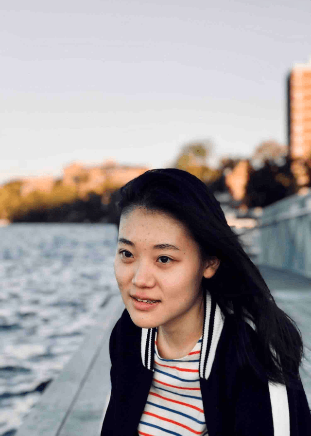 The image shows a young woman with long dark hair smiling at the camera, with a body of water and a cityscape in the background.