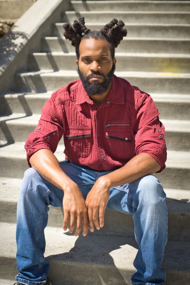 A man with dreadlocks wearing a red shirt and jeans sits on a set of concrete stairs in an outdoor setting.