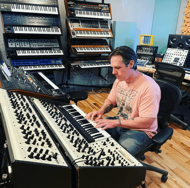 A man sits at a mixing console surrounded by various electronic musical instruments and equipment in a recording studio setting.