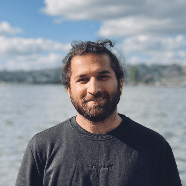 A smiling man with a beard stands in the foreground, with a body of water and cloudy sky visible in the background.
