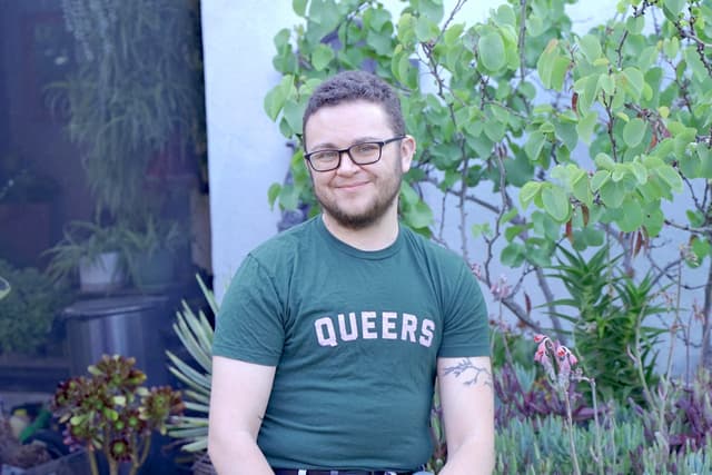 A smiling person wearing a green t-shirt with the word "QUEERS" printed on it, standing in front of a lush, green background with plants and foliage.