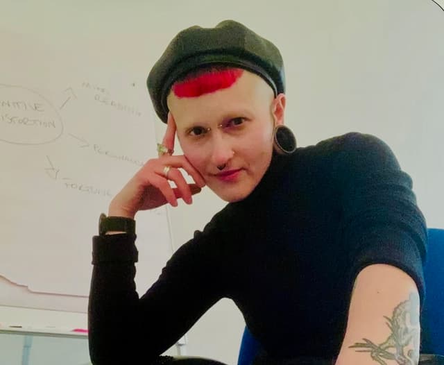 The image shows a person with short dark hair wearing a black hat and a black shirt, standing in front of a whiteboard with writing on it.