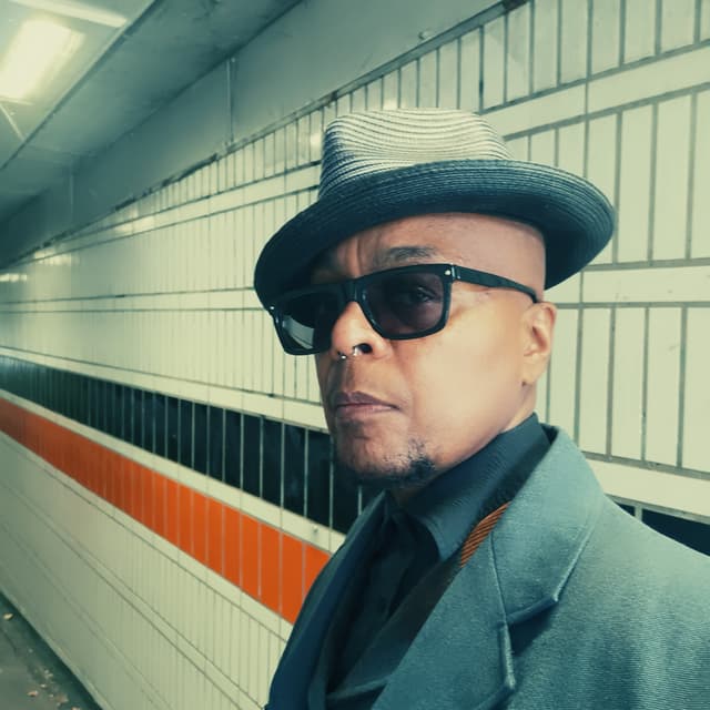 A man wearing a hat and sunglasses stands in front of a tiled wall with orange and white stripes.