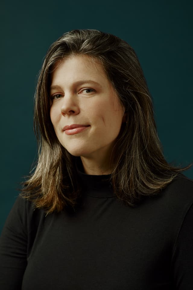 The image shows a woman with long, dark hair wearing a black turtleneck sweater against a dark green background.