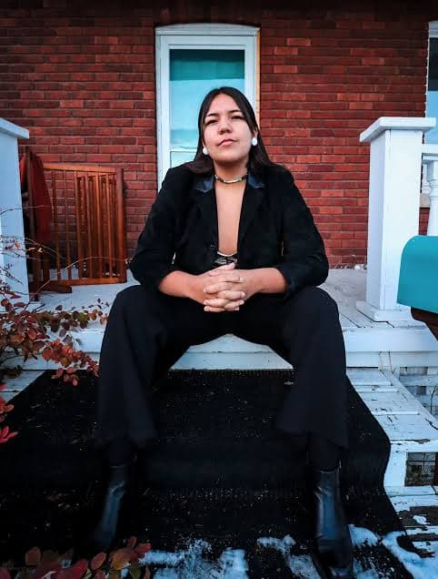 A young woman with dark hair sits on a wooden bench in front of a brick building, wearing a black coat and smiling at the camera.