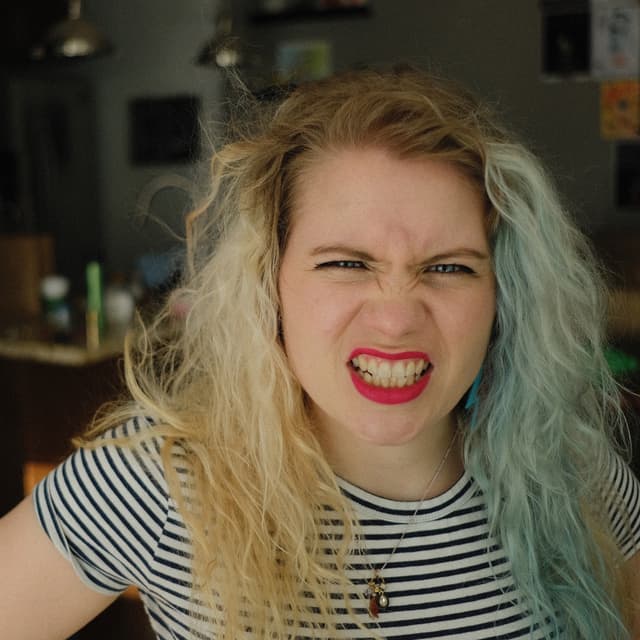 A woman with blonde and blue hair is making a fierce facial expression, her mouth open in a wide, toothy grin, against a dark background.