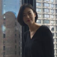 A young woman with dark hair stands in front of a window, gazing out at the cityscape visible through the glass.