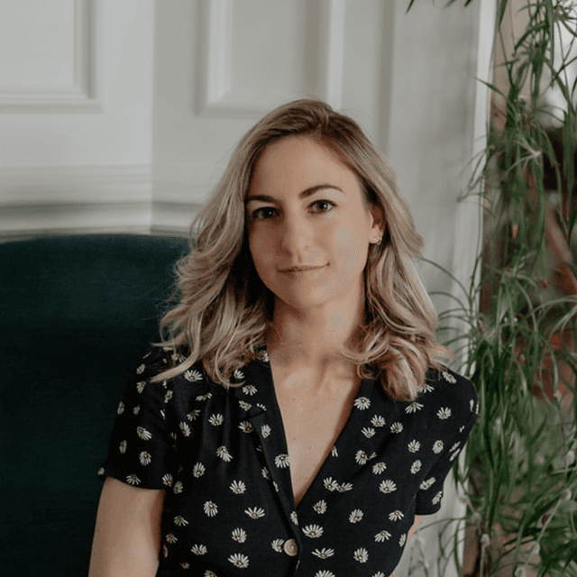 A young woman with wavy blonde hair sits in a green armchair, surrounded by potted plants in a cozy, indoor setting.