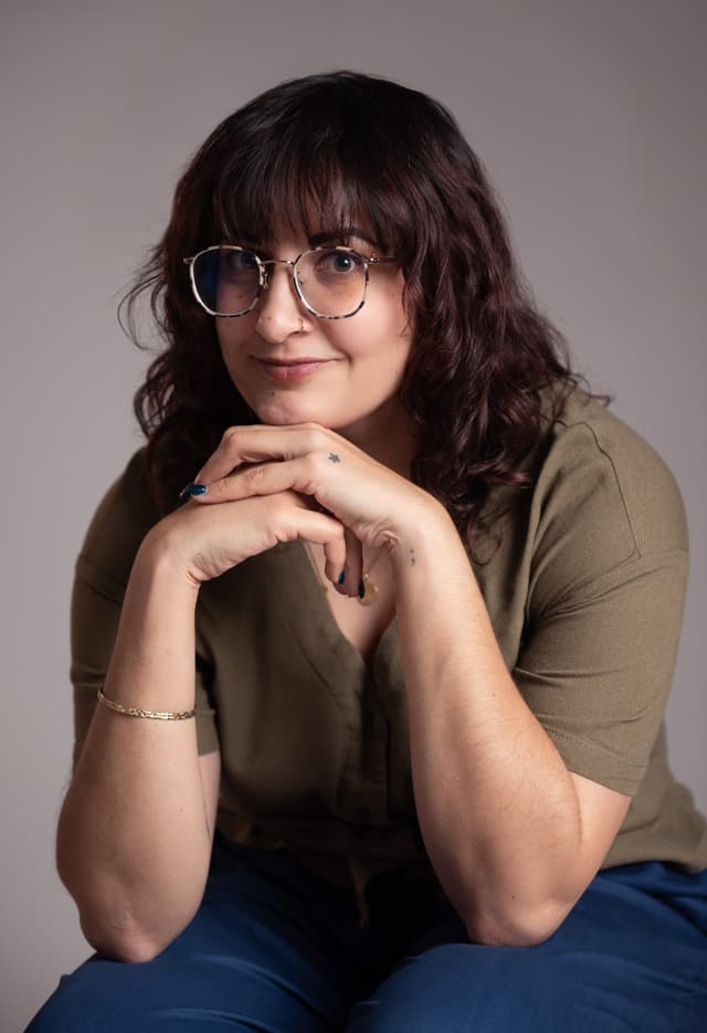 A person with vibrant purple hair sits cross-legged on a rocky shore, surrounded by a serene lake and mountains in the distance under an overcast sky.