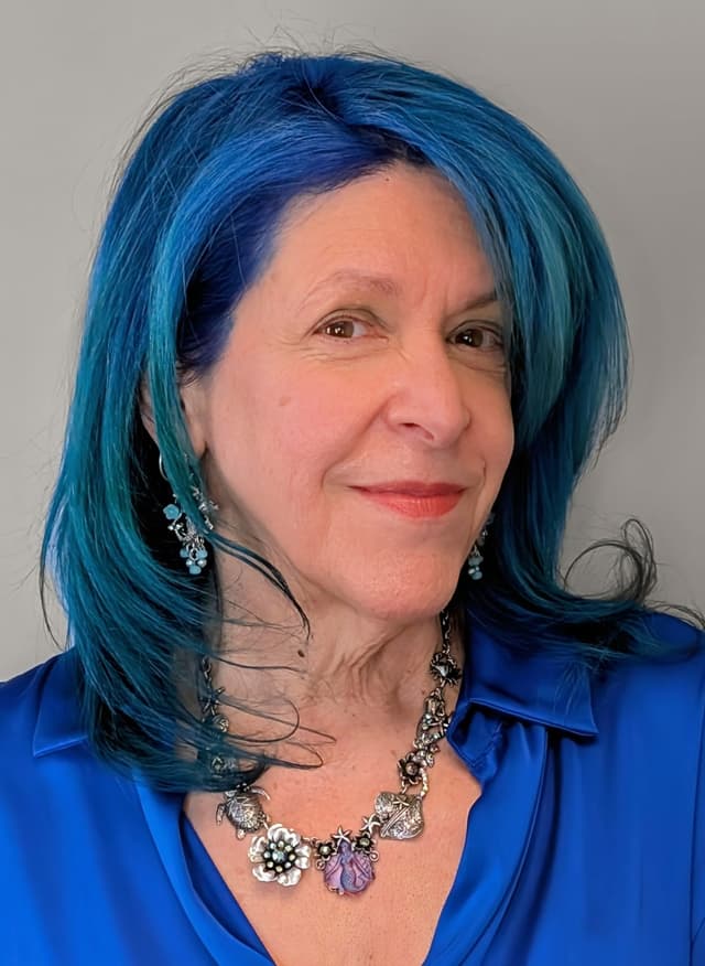 The image shows a woman with vibrant blue hair, wearing a blue blouse and an elaborate necklace, smiling warmly against a plain background.