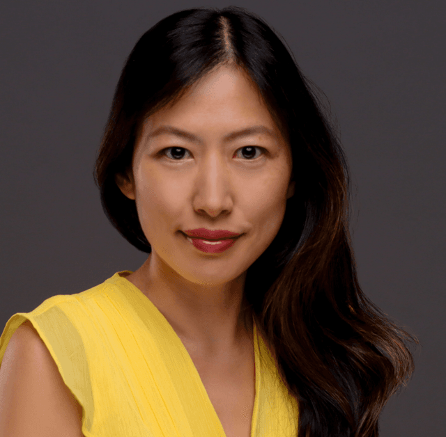 A woman with long dark hair wearing a yellow top is smiling against a dark background.