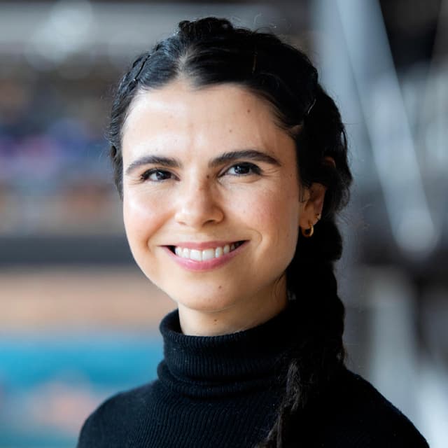 A smiling woman with dark hair in braids, wearing a black turtleneck sweater, stands in front of a blurred background.