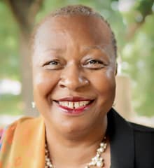 A smiling older woman with gray hair wearing a black jacket and pearls, set against a blurred natural background.