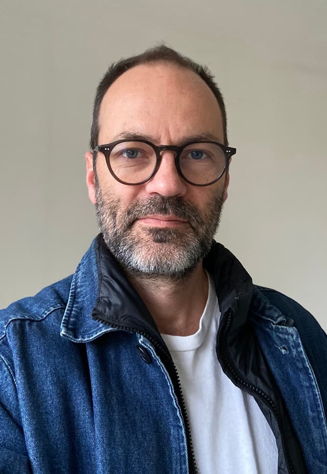 A middle-aged man with a beard and glasses, wearing a denim jacket, stands against a plain background.