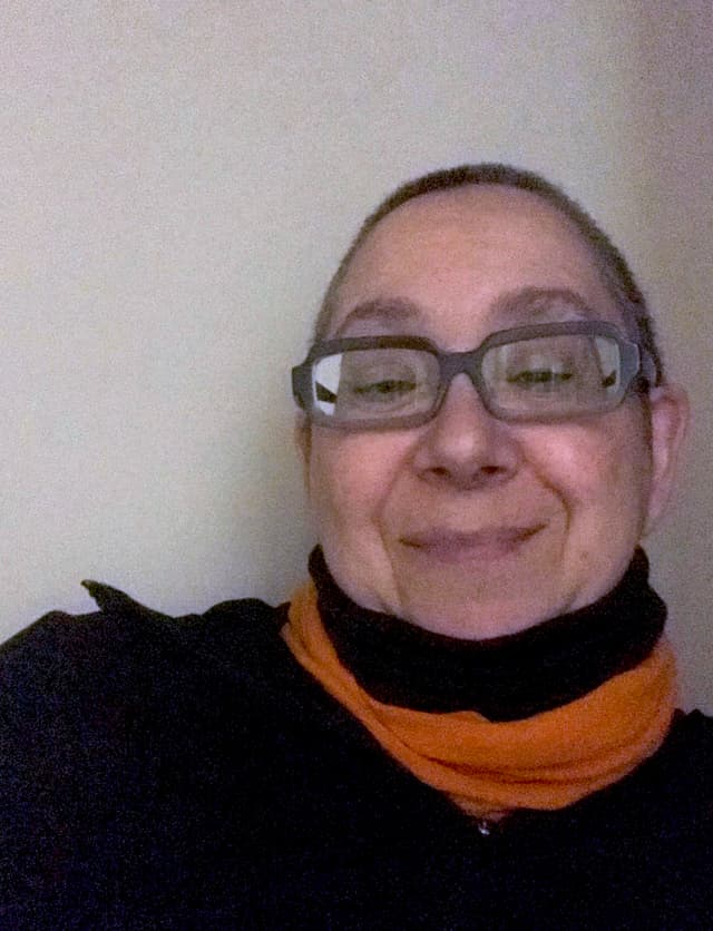 A smiling person wearing glasses and a black jacket with an orange scarf against a plain background.