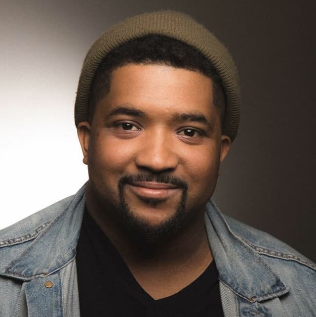 A smiling man with a beard wearing a beige knit hat and a denim jacket against a dark background.