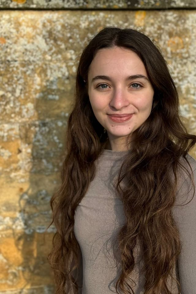 The image shows a young woman with long, wavy brown hair standing in front of a stone wall, smiling warmly at the camera.