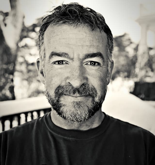 A middle-aged man with a beard and tousled hair gazes directly at the camera, set against a blurred outdoor background.