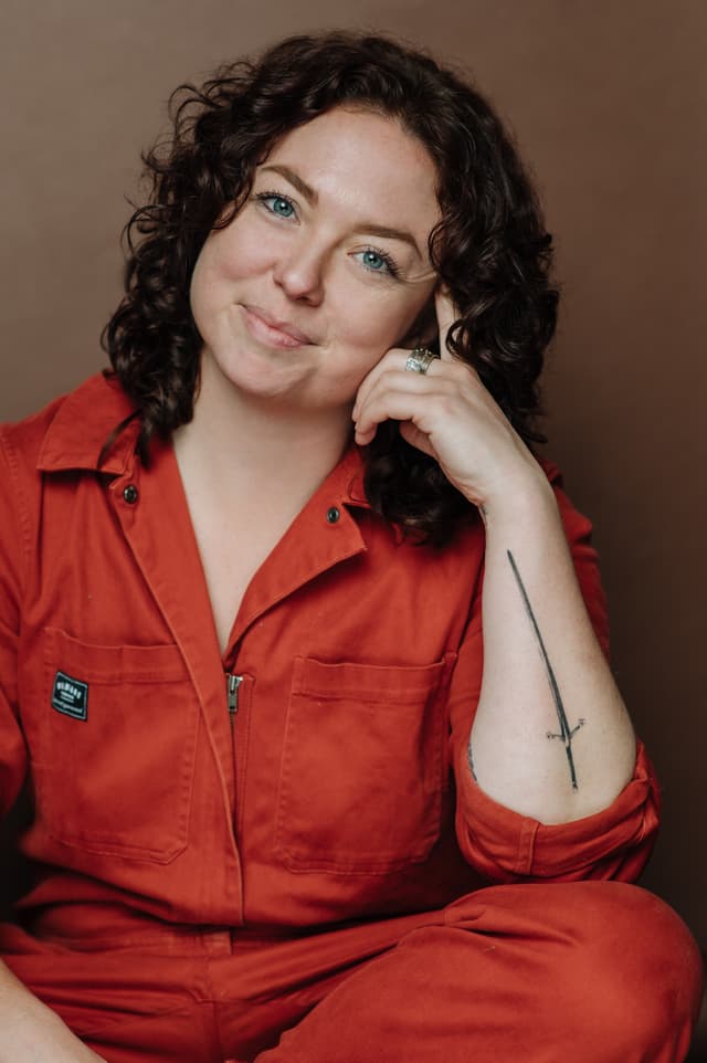 A woman with curly dark hair wearing a bright red jacket is smiling at the camera against a plain background.