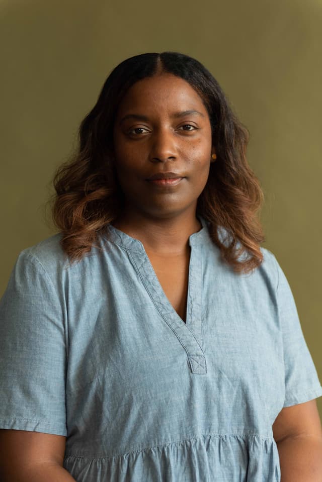 A woman with curly dark hair wearing a light blue blouse stands against a neutral background.