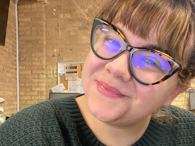 A woman with glasses and a sweater is smiling at the camera in what appears to be an office or workplace setting.