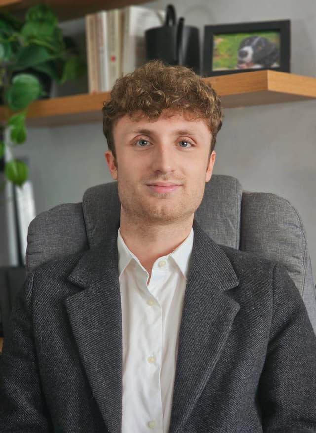A young man with curly hair wearing a gray suit jacket and a white shirt is sitting in an office setting with shelves and plants visible in the background.