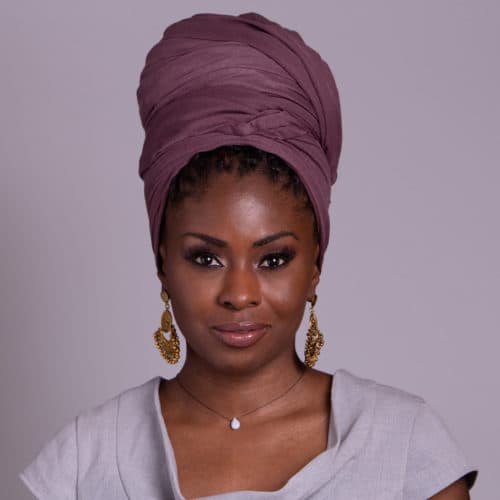 The image depicts a woman with dark skin and curly hair wearing a burgundy turban-style headpiece, gold hoop earrings, and a white top against a plain purple background.