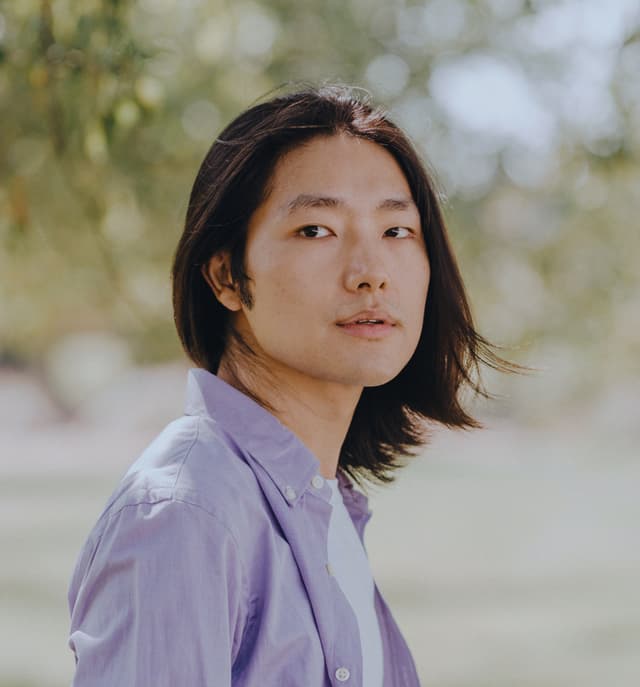 The image shows a young woman with long dark hair wearing a light purple shirt, standing in a natural outdoor setting with blurred foliage in the background.