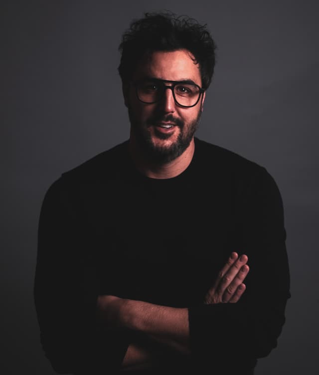 A man with a beard and glasses, wearing a black shirt, stands in a dark background.