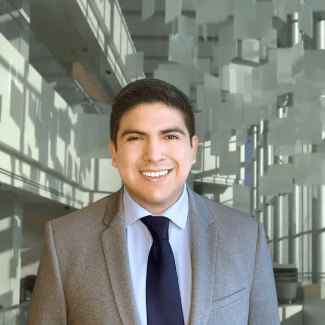 A smiling young man in a gray suit stands in front of a modern glass and steel building.