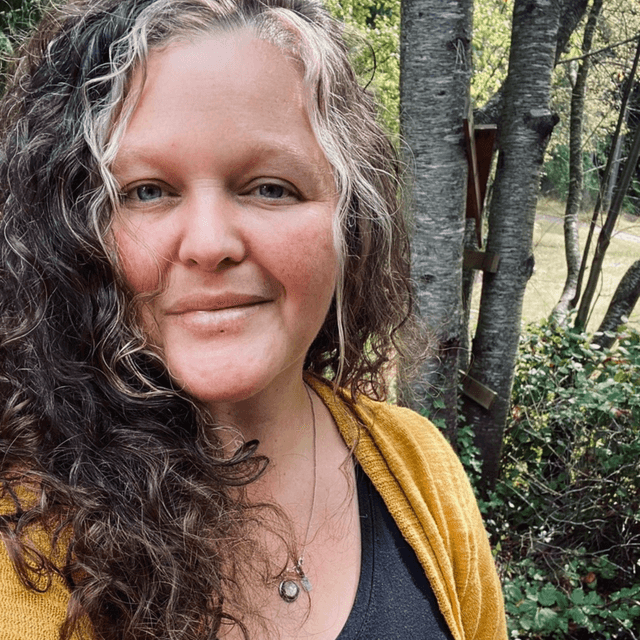 A woman with curly, shoulder-length hair stands in a forested area, wearing a yellow cardigan and a necklace, with a serene expression on her face.
