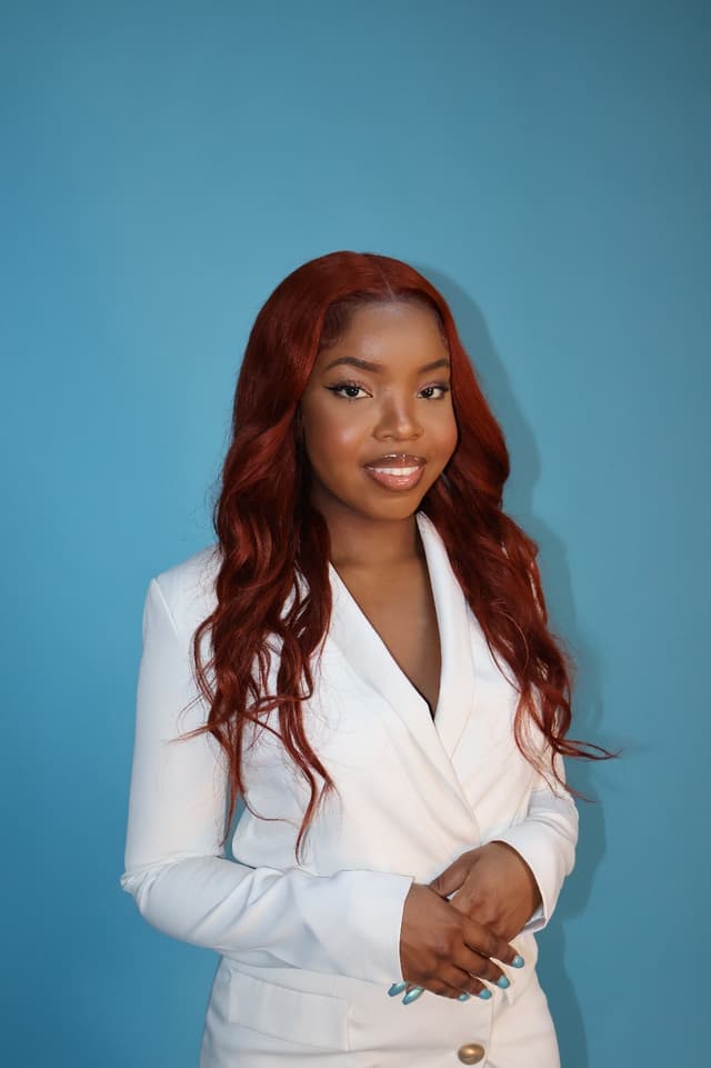 A young woman with long, wavy red hair stands in a white blazer against a vibrant blue background.