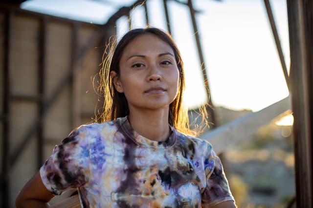 A young woman with long dark hair stands in front of a wooden structure, her face illuminated by the warm glow of the setting sun.