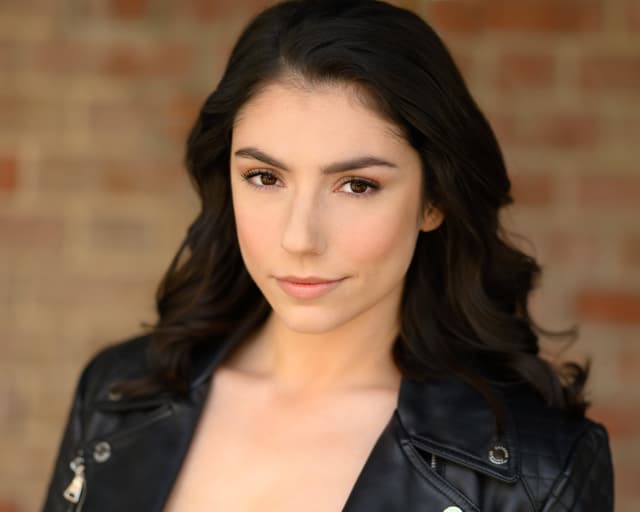 A young woman with dark hair and a serious expression, wearing a black leather jacket, stands in front of a brick wall background.