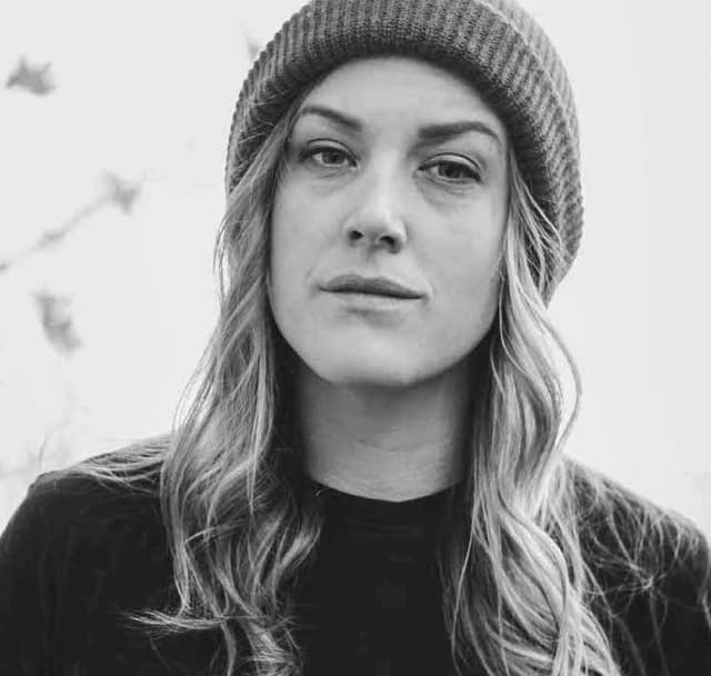 A woman with long, wavy blonde hair wearing a gray knit hat and a black sweater, set against a blurred natural background.