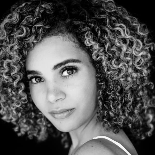 The image depicts a close-up portrait of a young woman with curly, dark hair and a thoughtful expression on her face. The background is dark, creating a dramatic contrast that draws the viewer's attention to the subject's features.