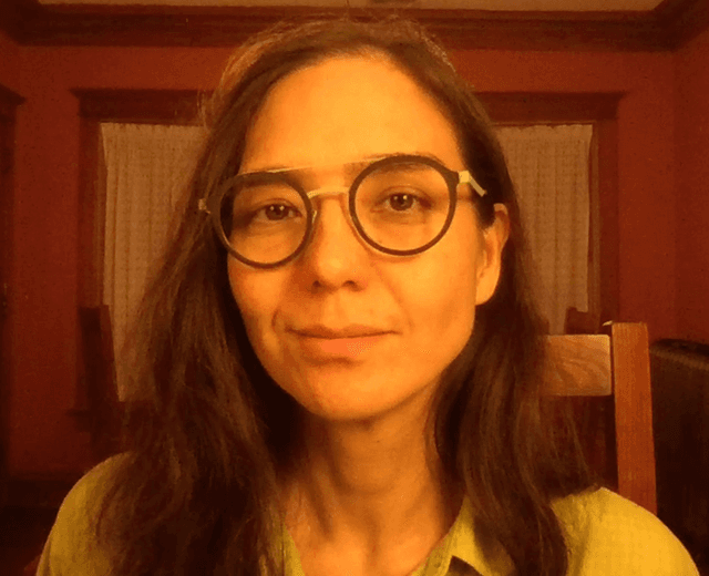 A person with long dark hair and glasses is shown in the foreground, with a wooden interior visible in the background.