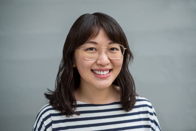A young woman with dark hair and glasses is smiling brightly against a plain gray background.