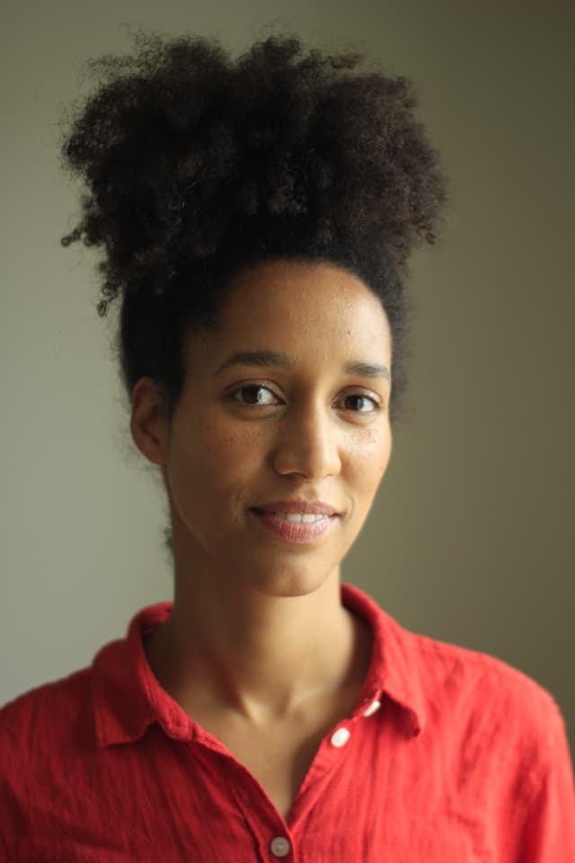 A young woman with curly dark hair wearing a red shirt smiles warmly against a neutral background.