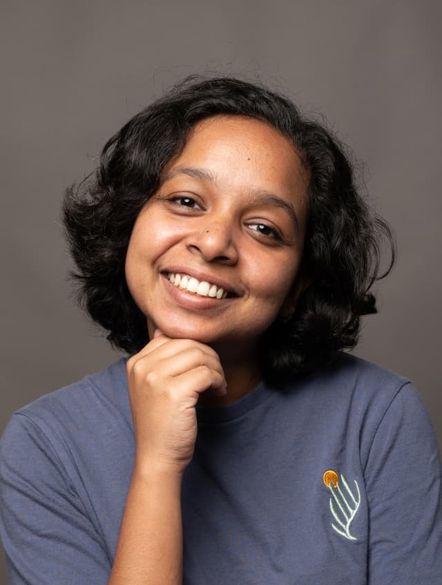 A smiling woman with curly dark hair wearing a gray shirt and resting her chin on her hand against a plain gray background.