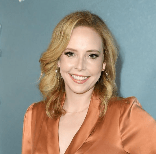 A smiling woman with wavy blonde hair wearing an orange dress stands in front of a gray background.