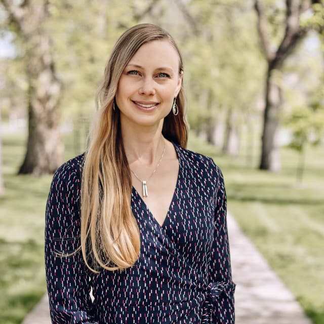 A smiling woman with long blonde hair stands in a park-like setting, surrounded by trees in bloom.
