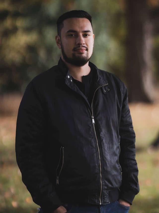 A man with a beard wearing a black jacket stands in a wooded outdoor setting.