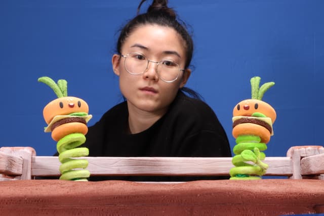 A young woman with glasses sits at a table, holding two colorful toy caterpillars in front of a vibrant blue background.