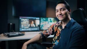 A smiling man sits at a desk in front of a computer monitor displaying various visual elements.