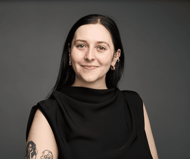 A woman with long dark hair, wearing a black top, smiling at the camera against a plain gray background.